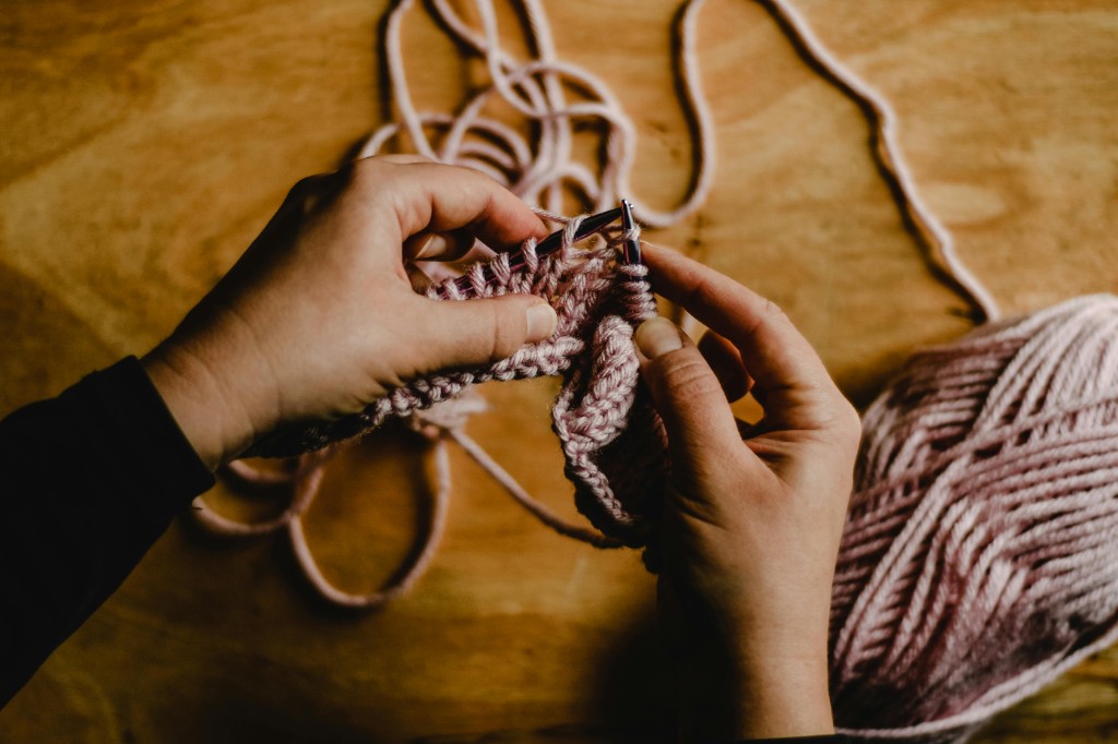 Hands knitting pink ribbed fabric with yarn and wooden needles on the table